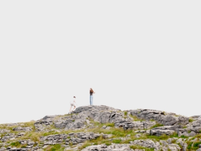 Three people—an adult and two children—standing on a rocky hillside, looking out over a grassy, rugged outcrop beneath a pale sky.