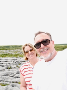Smiling couple taking a selfie outdoors on a rocky field with green hills in the background.