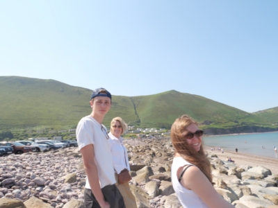 Group of people standing on a rocky beach with green hills and a clear blue sky behind them.