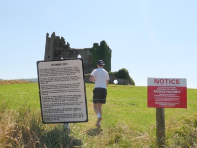 Ruined stone castle with tall, crumbling walls behind informational signs in a grassy field under a blue sky.