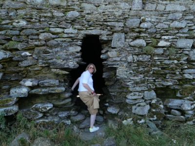 Woman in a white shirt and tan shorts stands at a dark stone doorway in a mossy, dry-stone wall ruin.