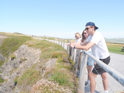 Three friends in white shirts lean on a wooden railing along a sunny hillside overlooking a rocky slope with wildflowers.