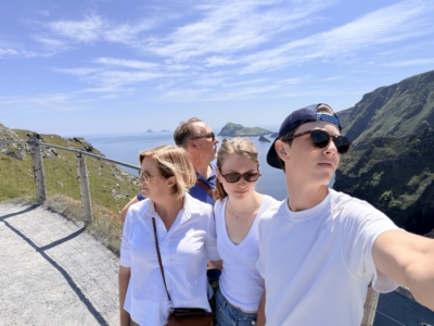 Group of people wearing white shirts posing for a selfie on a cliff with a coastal landscape, blue sky, and sea in the background.