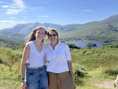 Two smiling women pose together outdoors against a backdrop of green hills, mountains, and a blue lake under a clear sky.