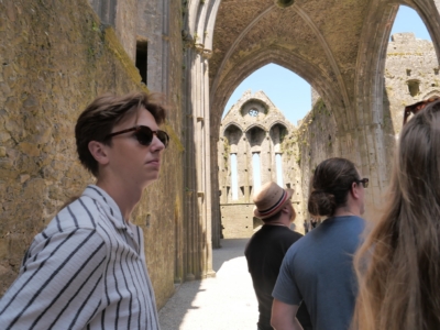Tourists stand among ancient stone ruins with tall Gothic arches and sunlit interior walls.
