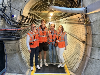 Four people in orange safety vests pose in a tunnel lined with cables and piping, standing on a yellow-edged grate floor.