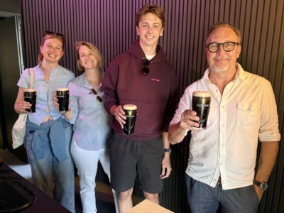 Four friends posing for a photo, each holding a Guinness pint and smiling at the camera in a bar with dark vertical paneling behind them.
