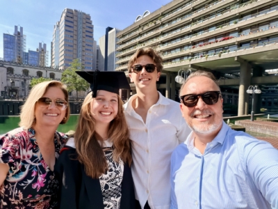 Group of five friends taking a sunny city selfie, smiling with modern buildings in the background; one person wearing a graduation cap and gown.