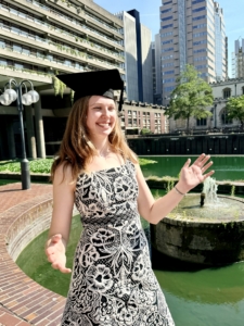 Smiling woman in a graduation cap and a patterned dress, arms raised, with a canal and modern buildings in the background.