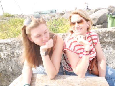 Two women sitting on a wooden bench by a rocky shoreline, smiling at the camera with sunglasses on in bright daylight.