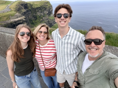 Four friends posing for a selfie on a cliff overlooking the ocean, with a grassy path and railing in the background.