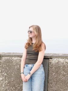 Young woman with long hair wearing sunglasses and a dark green tank top, standing by a stone wall near the water, smiling slightly.