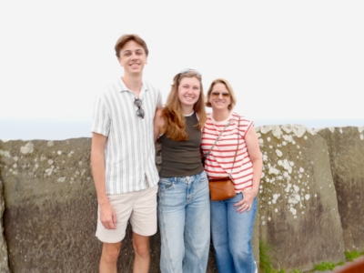 Three people pose together outdoors against a stone wall with the sea in the background, smiling at the camera.