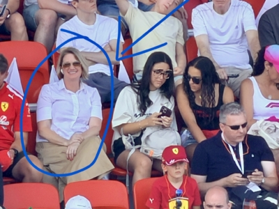 Smiling woman in a white blouse and beige skirt with sunglasses seated in red stadium seats among spectators.