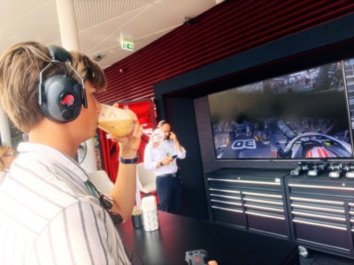 Person wearing over-ear headphones sipping an orange beverage while a racing video plays on a large screen nearby.