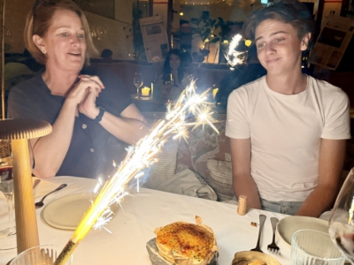 Two people sit at a restaurant table with a bright sparkler in the foreground; woman on the left smiles, man on the right wears a white shirt and smiles back, birthday vibe likely.