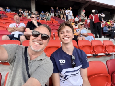 Two men take a selfie in a stadium with bright red seats and a crowd in the background, smiling.