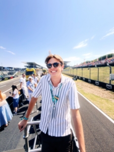 Smiling man in a striped shirt and sunglasses at a race track, with spectators in the background.