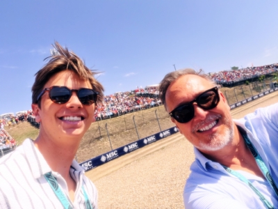 Two people take a sunny outdoor selfie, wearing sunglasses with a large crowd behind them at a sporting event or festival.