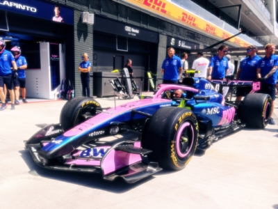 Pit crew in blue shirts surrounding a pink-blue race car in the pit lane at a racing event.