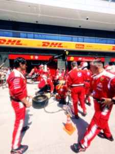Ferrari pit crew in red suits gathered in the pit lane with tires and equipment visible, DHL signs in the background