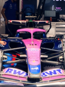 Front-on view of a pink-and-blue BWT Alpine Formula 1 car in the garage, with a technician in a blue shirt nearby.