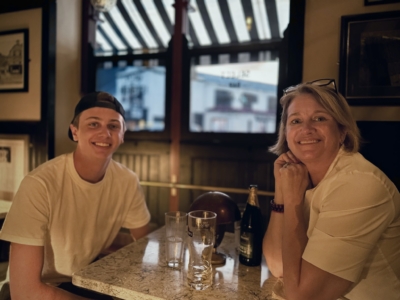 Two people smiling at a marble-topped bar, woman on the right with glasses on her head and a man on the left wearing a cap.