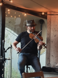 Musician wearing a hat playing a violin on a small stage with warm string lights above him