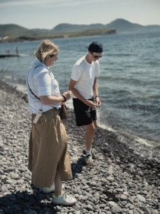 Two people walk along a rocky shoreline by calm water, one woman in a white shirt and tan skirt, a man in a white t-shirt and cap beside her.