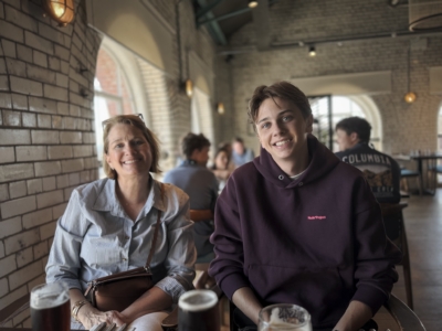 Two people sit at a cafe table smiling, drinks in front of them, brick walls and warm lighting in the background.