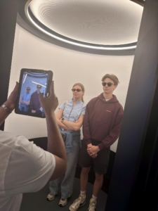Three teenagers posing for a photo in a white exhibit space, wearing sunglasses, while someone uses a tablet to capture their reflection.